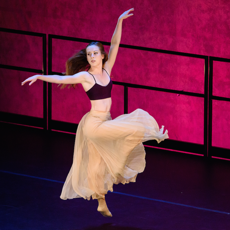 A female dancer from Bugge Ballet leaps into a front attitude position, her hair lofts in the air 